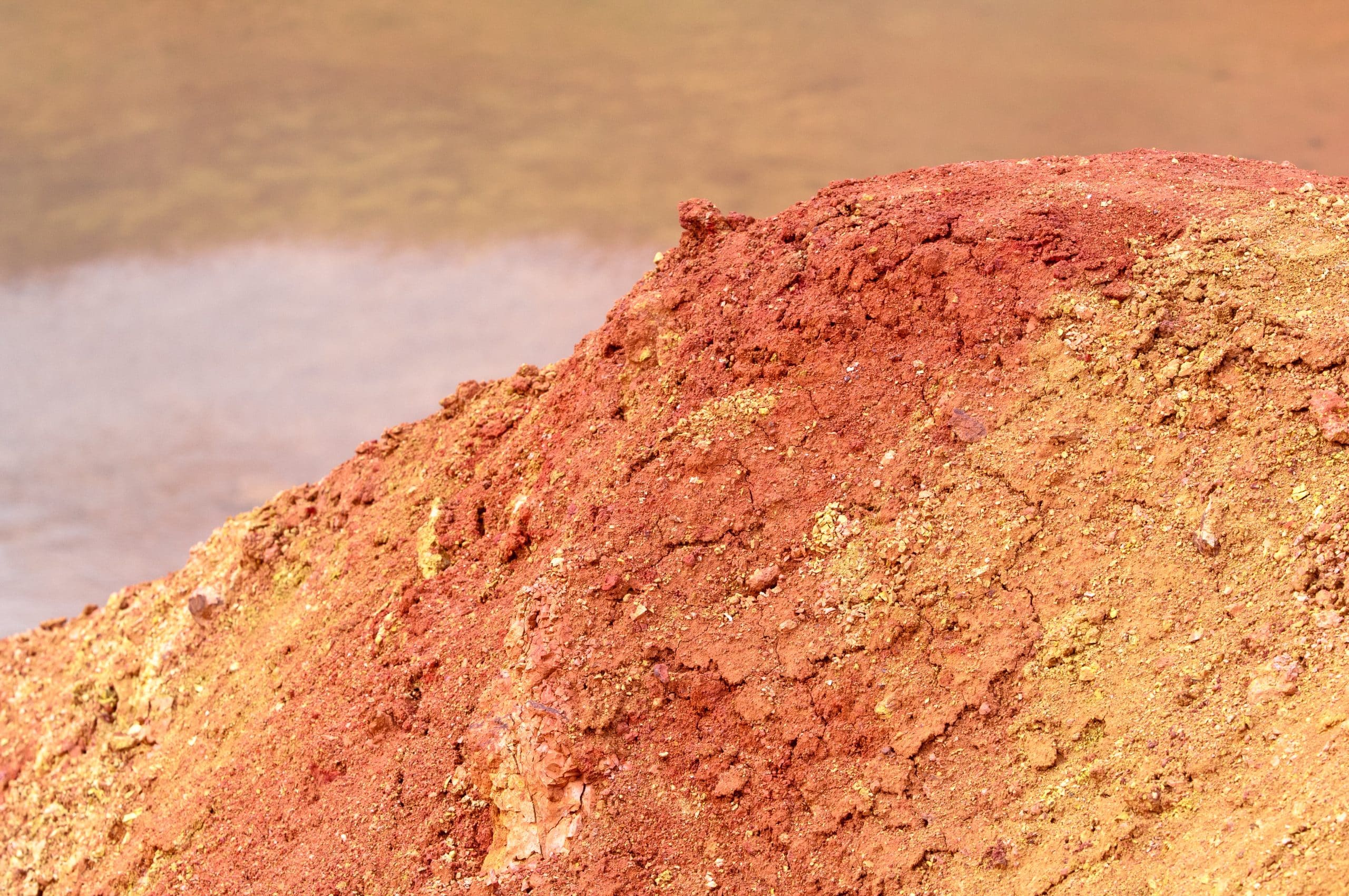 A close-up of a mound of red and yellow earth with a rough, textured surface, possibly including spent pot lining. The background is blurred, showing a body of muddy water, creating a natural contrast between the bright, dry soil and the wet, muted surroundings.