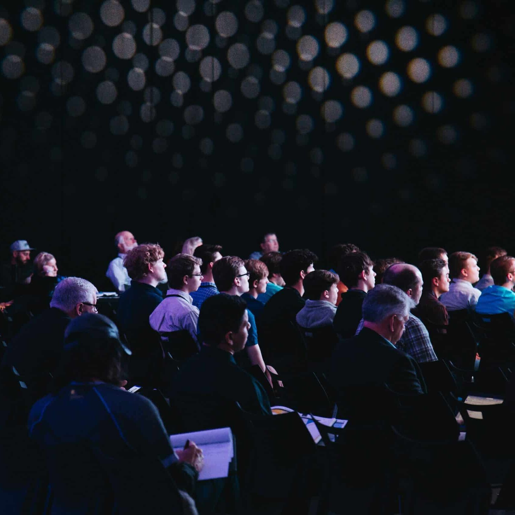 Audience members seated and paying attention to a presentation in a dimly lit room with dotted lights overhead, creating a focused and professional atmosphere, as the press quietly takes notes.