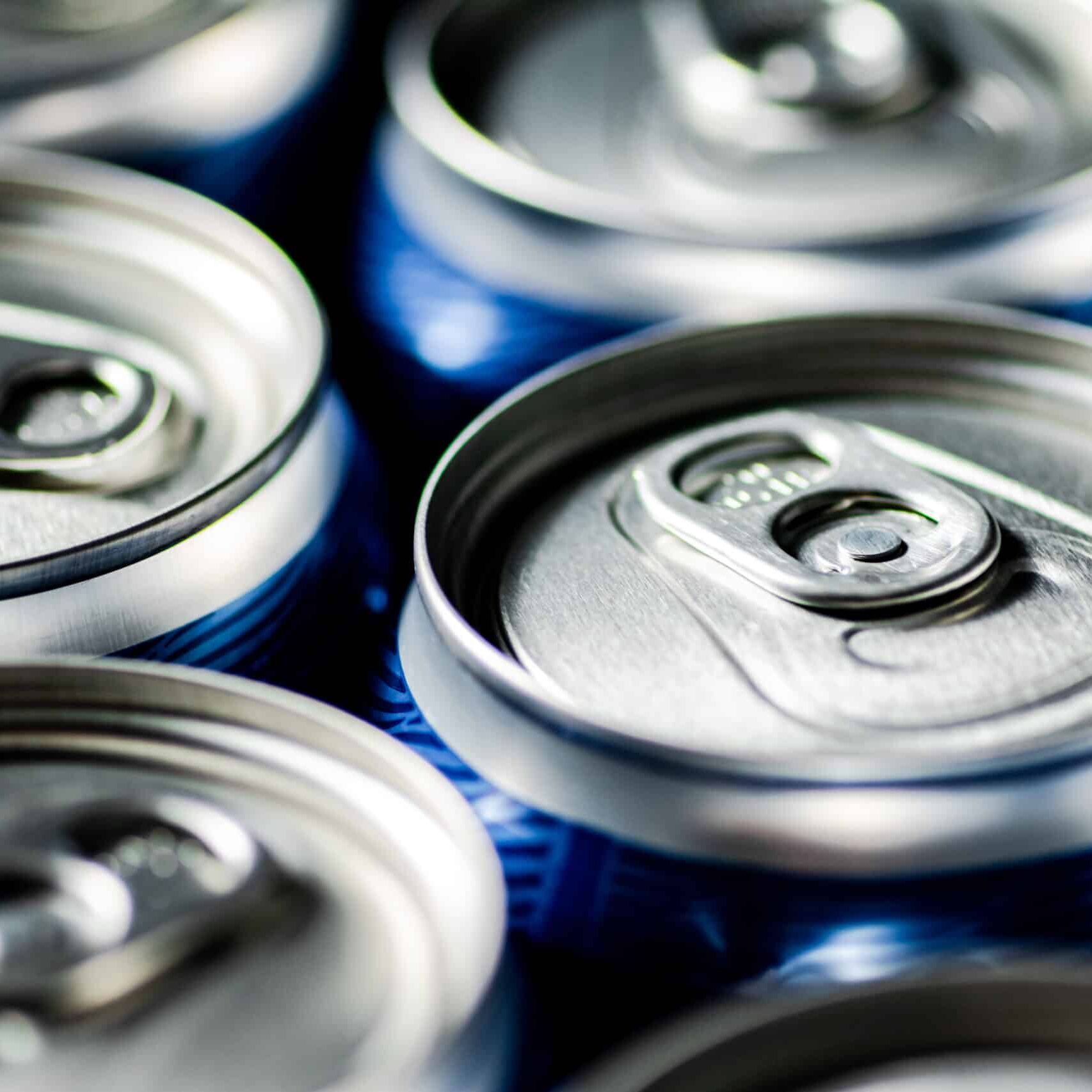 Close-up view of several aluminium soda cans with pull-tab openings. The cans are tightly packed together and have a blue design along the sides, though only the tops are in clear focus, highlighting their sleek look amidst various environmental claims.