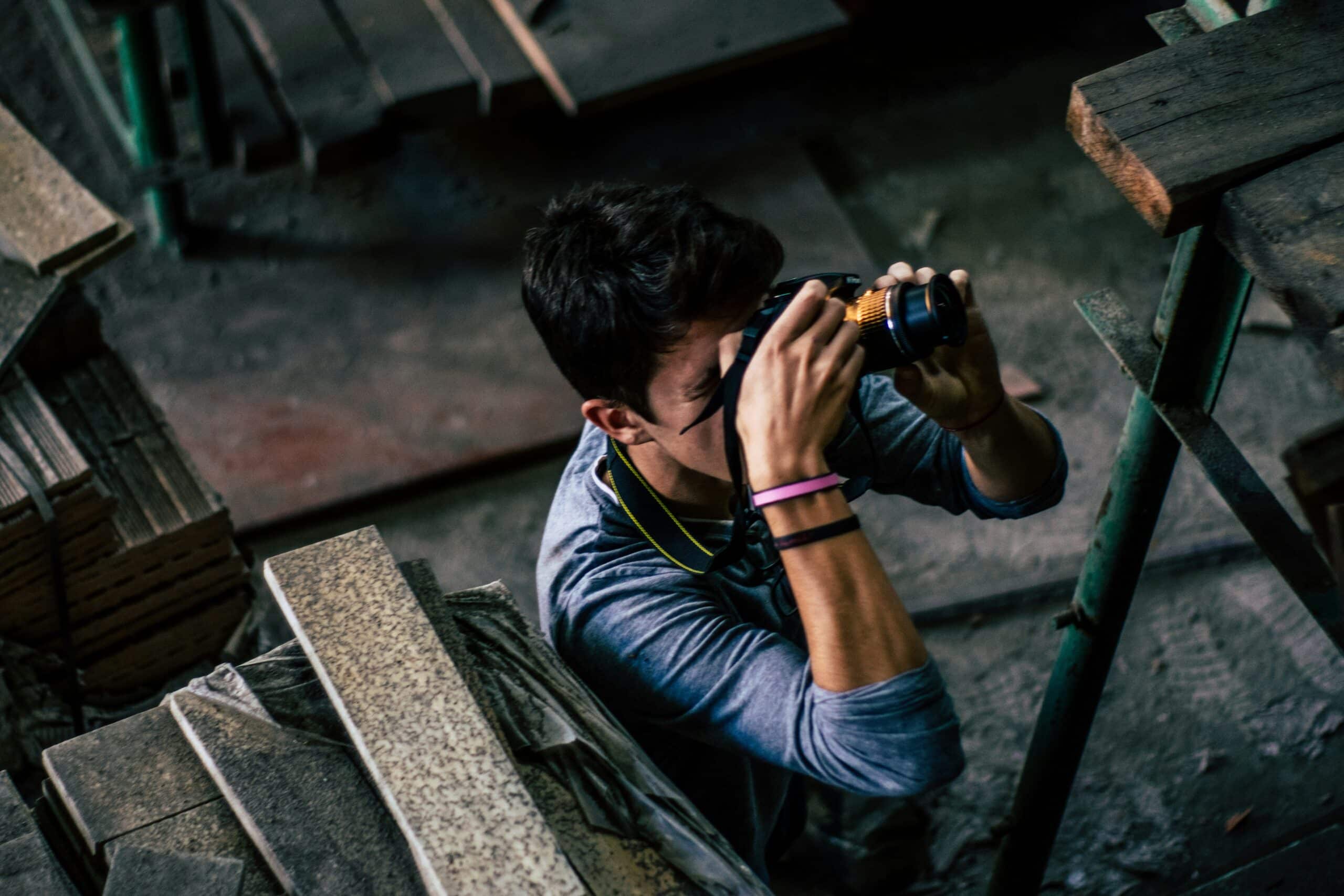 A person with short dark hair is holding a camera up to their eye, capturing a photo in an environment filled with construction materials and wooden planks. They are wearing a long-sleeved shirt and a wristband. The scene appears cluttered and industrial, resembling a press photographer's workspace.