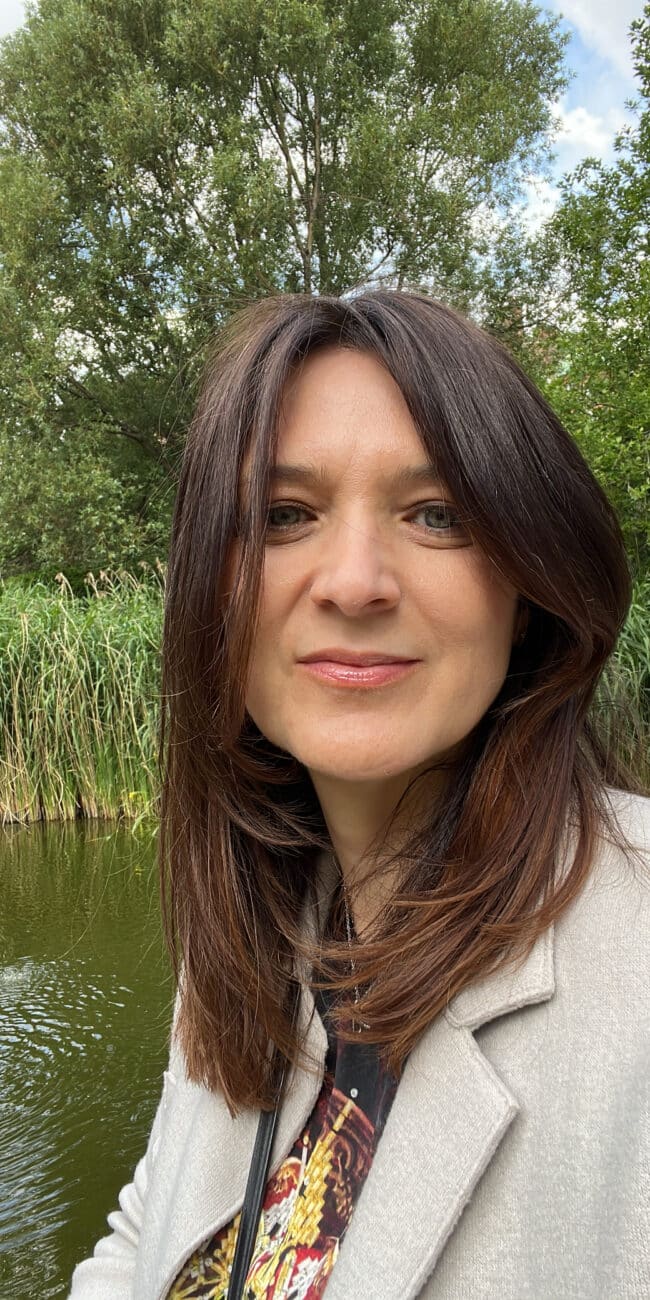 A woman with straight brown hair and a light jacket stands outdoors near a pond, perhaps reflecting after a team outing, with tall green reeds and trees in the background.