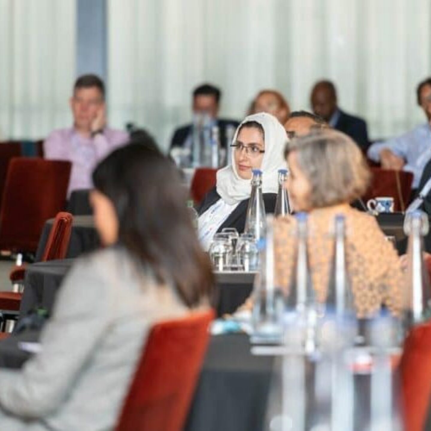 A diverse group of people is seated in the conference room for the 29th CRU, with tables arranged in rows. Attendees appear to be listening attentively to a speaker. Some are taking notes while others have beverages in front of them. The room is well-lit with large windows.