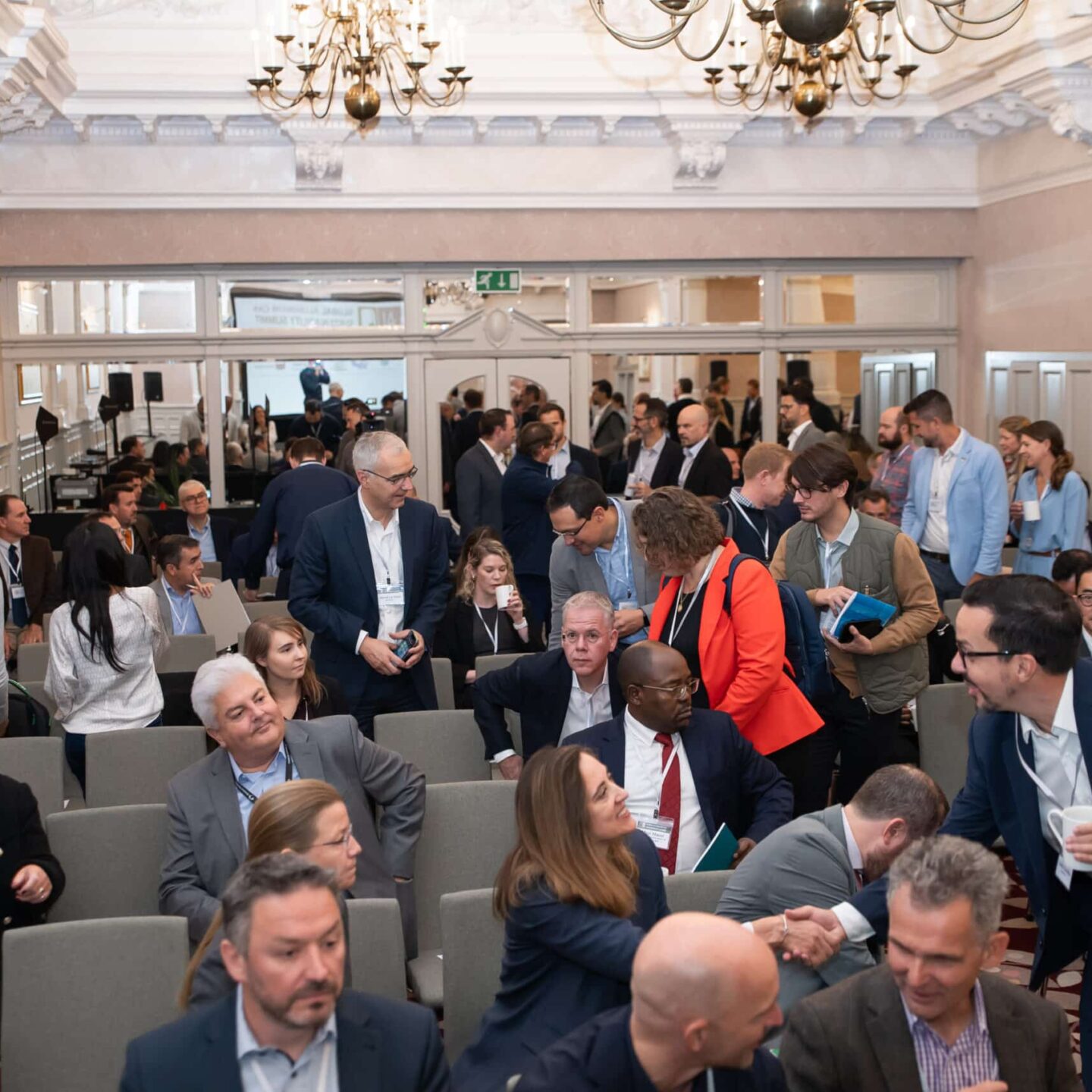A group of people in business attire are gathered in a conference room adorned with chandeliers, engaging in conversations. Among them are representatives from the International Aluminium Institute, all eagerly waiting for the event to begin.