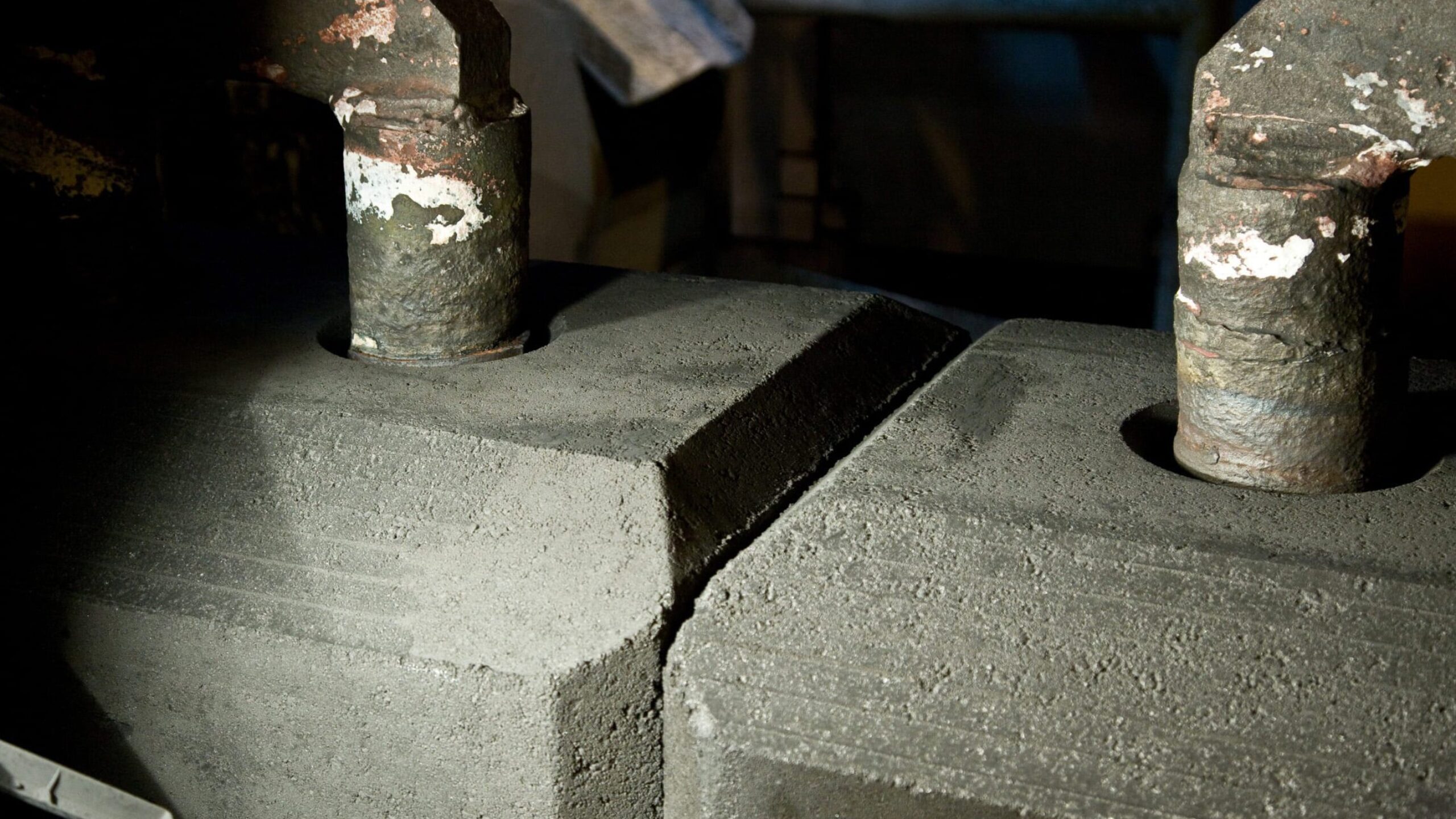 Close-up of industrial equipment with two metal pipes extending into a rectangular brick-like structure covered in dark, dusty material. The surrounding area is dimly lit, highlighting the texture and wear from aluminium processing on the pipes and surface where fluorides are often present.