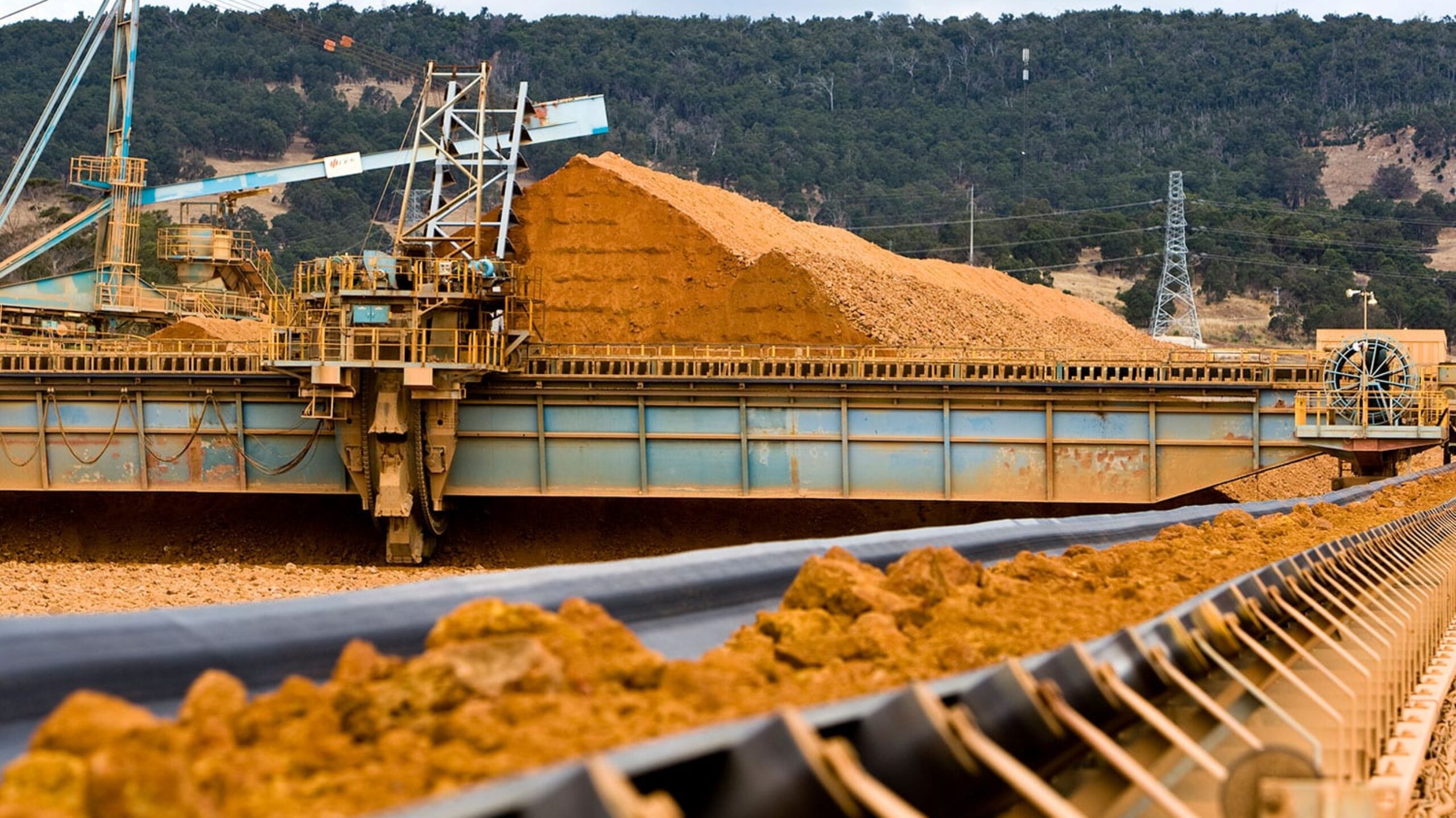 A mining site with machinery processing large amounts of earth material. Conveyor belts transport the mined material, and a large pile of excavated earth is visible in the background. The International Aluminium Institute oversees operations here. Hilly terrain and forested areas are in the distance under a cloudy sky.