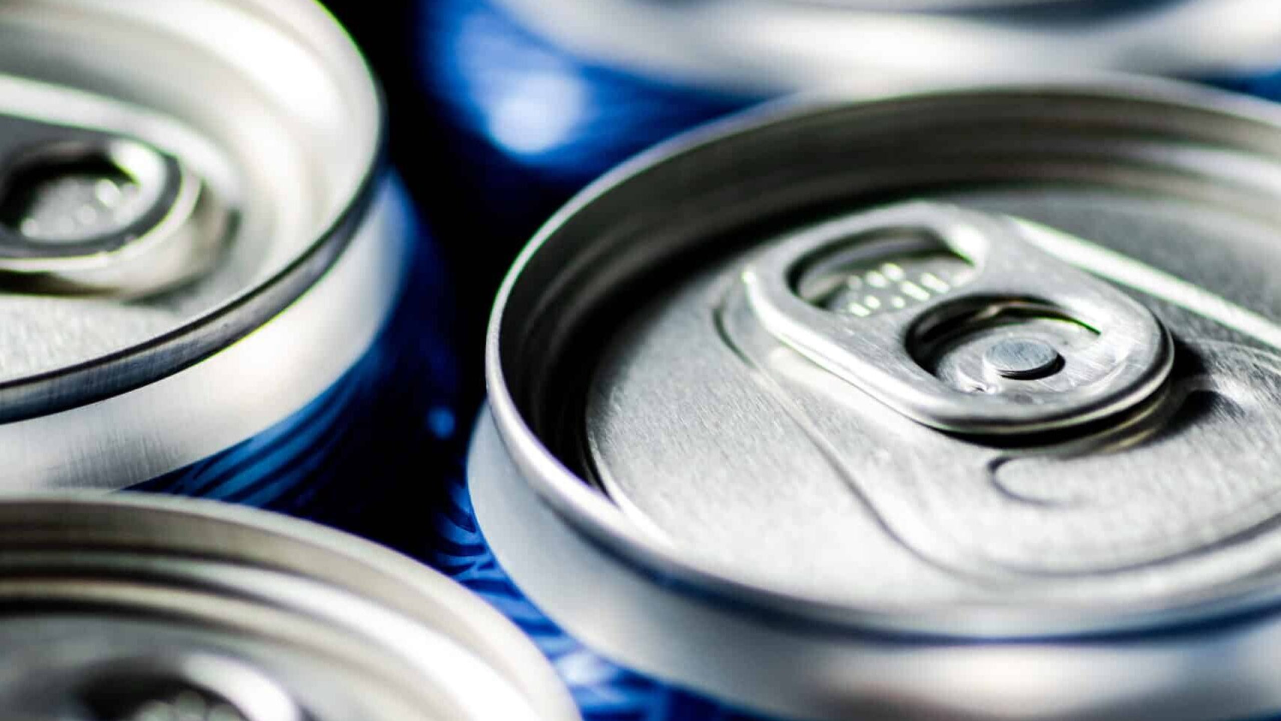 Close-up view of several aluminium soda cans with pull-tab openings. The cans are tightly packed together and have a blue design along the sides, though only the tops are in clear focus, highlighting their sleek look amidst various environmental claims.