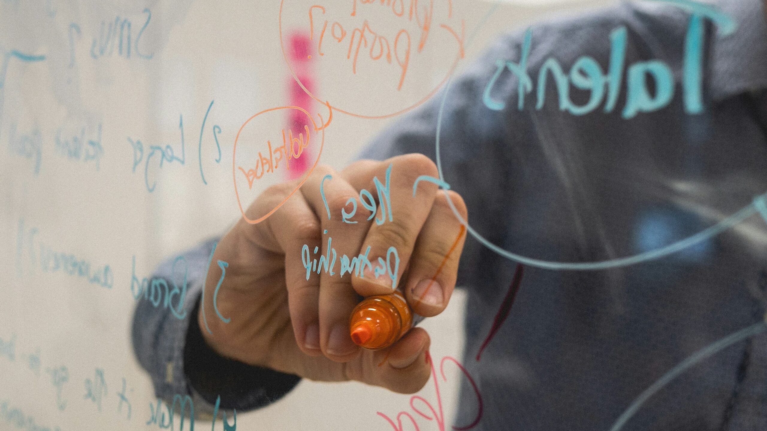A person is writing on a transparent glass board with a red marker. The board has various notes, keywords, and diagrams in different colors, including the words “key,” “relationship,” and "aluminium 2030." The person is wearing a patterned, long-sleeve shirt.