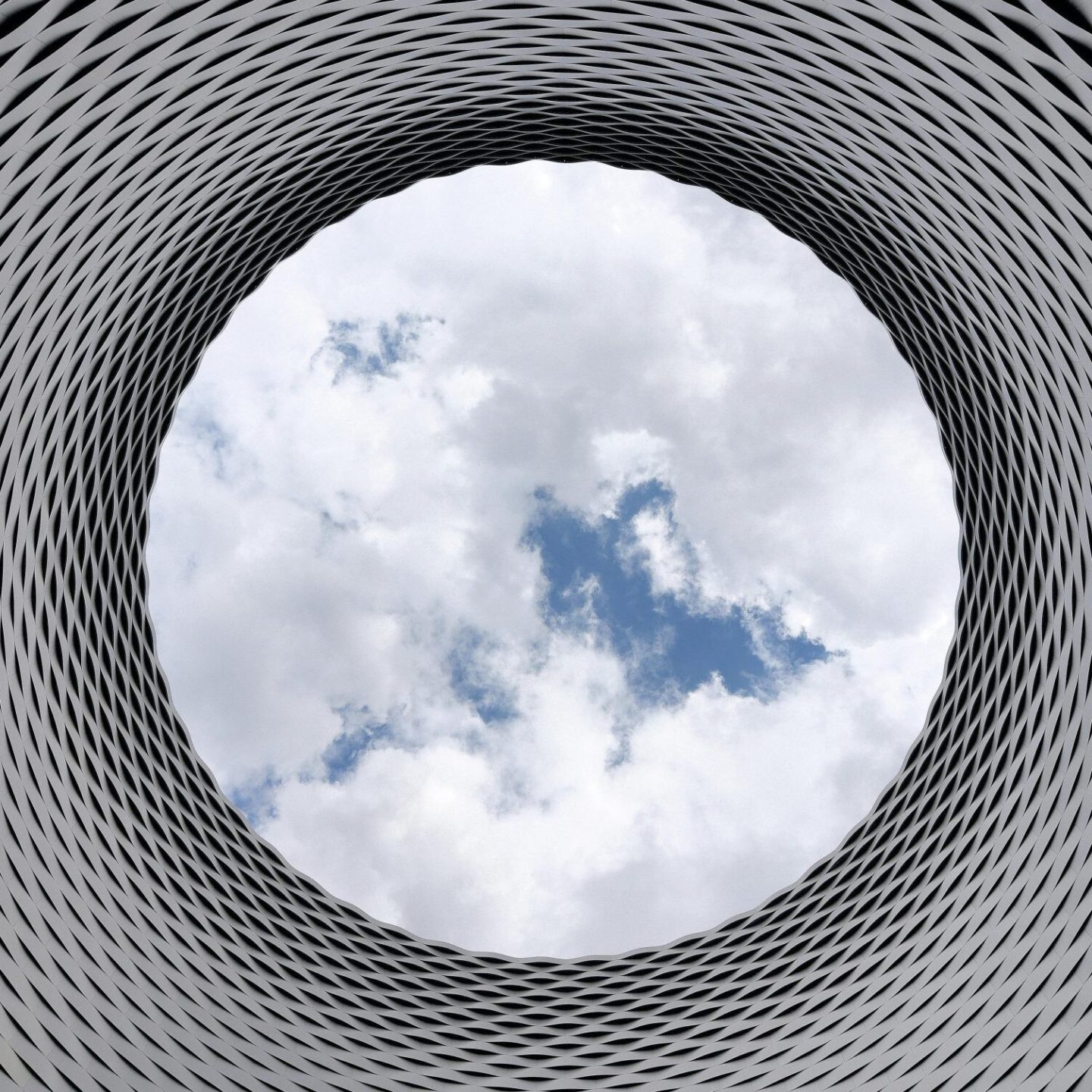 A view of a circular opening in a modern architectural structure with a geometric, grid-like pattern celebrates the IAI 50 anniversary. Through the opening, a partly cloudy sky is visible, creating a stark contrast between the structured pattern of the building and the natural sky.
