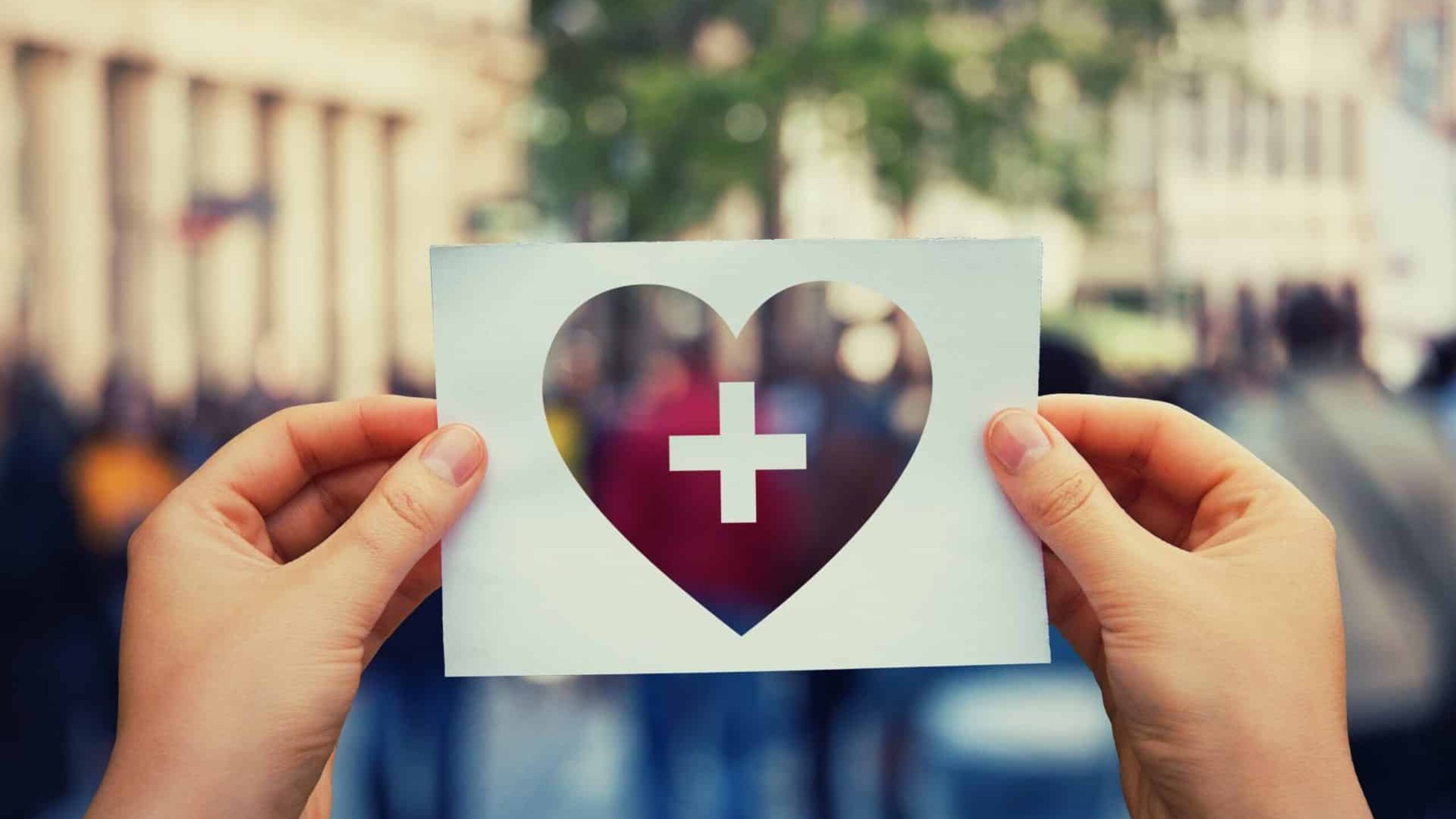 Two hands hold up a piece of paper with a heart shape cutout containing a white cross, against a blurred urban background with people and buildings. The paper is held at the center of the image, emphasizing the heart and cross symbol, highlighting a powerful message about consumer health.