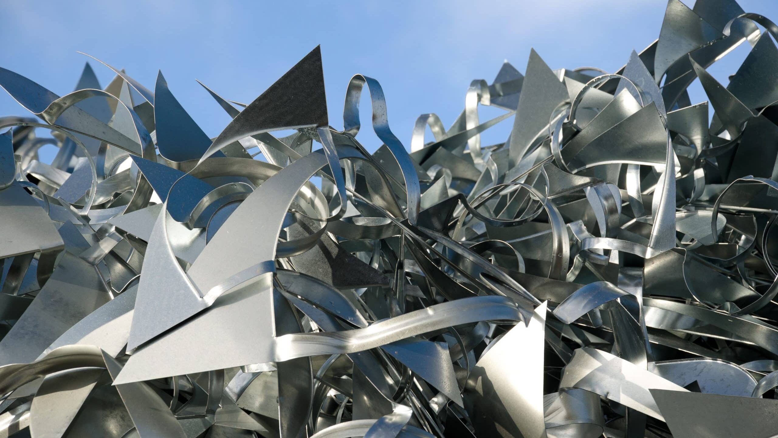 A pile of sharp, angular aluminium scrap with various shapes and edges is heaped against a backdrop of a clear blue sky. The metallic pieces reflect light, creating a shiny, contrasting surface.