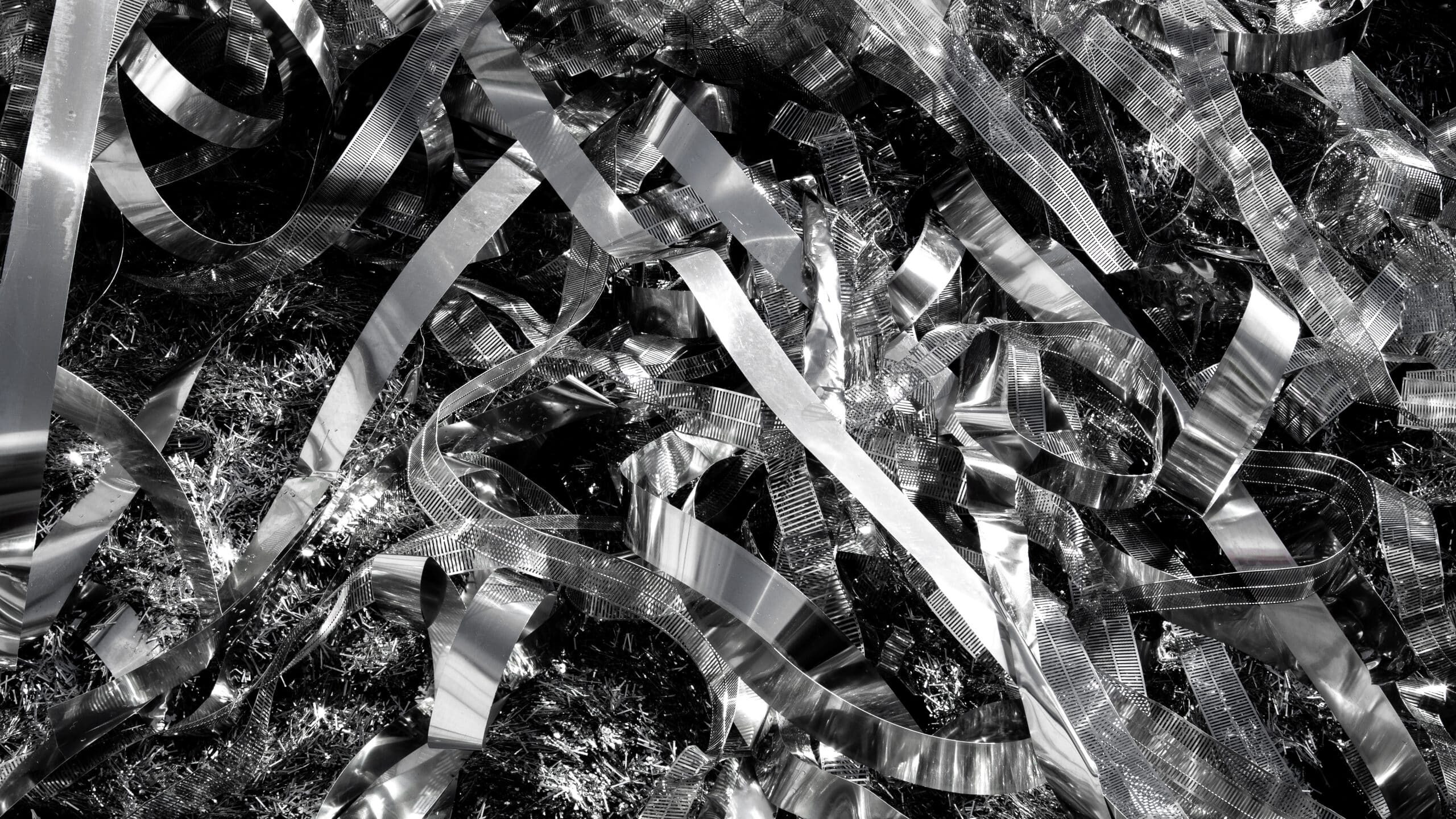 A black-and-white image depicts a chaotic arrangement of reflective, metallic ribbons strewn across a rough-textured surface, possibly grass. The shiny aluminium ribbons catch and reflect light, creating a striking contrast with the darker background, hinting at their lifecycle amidst nature's elements.