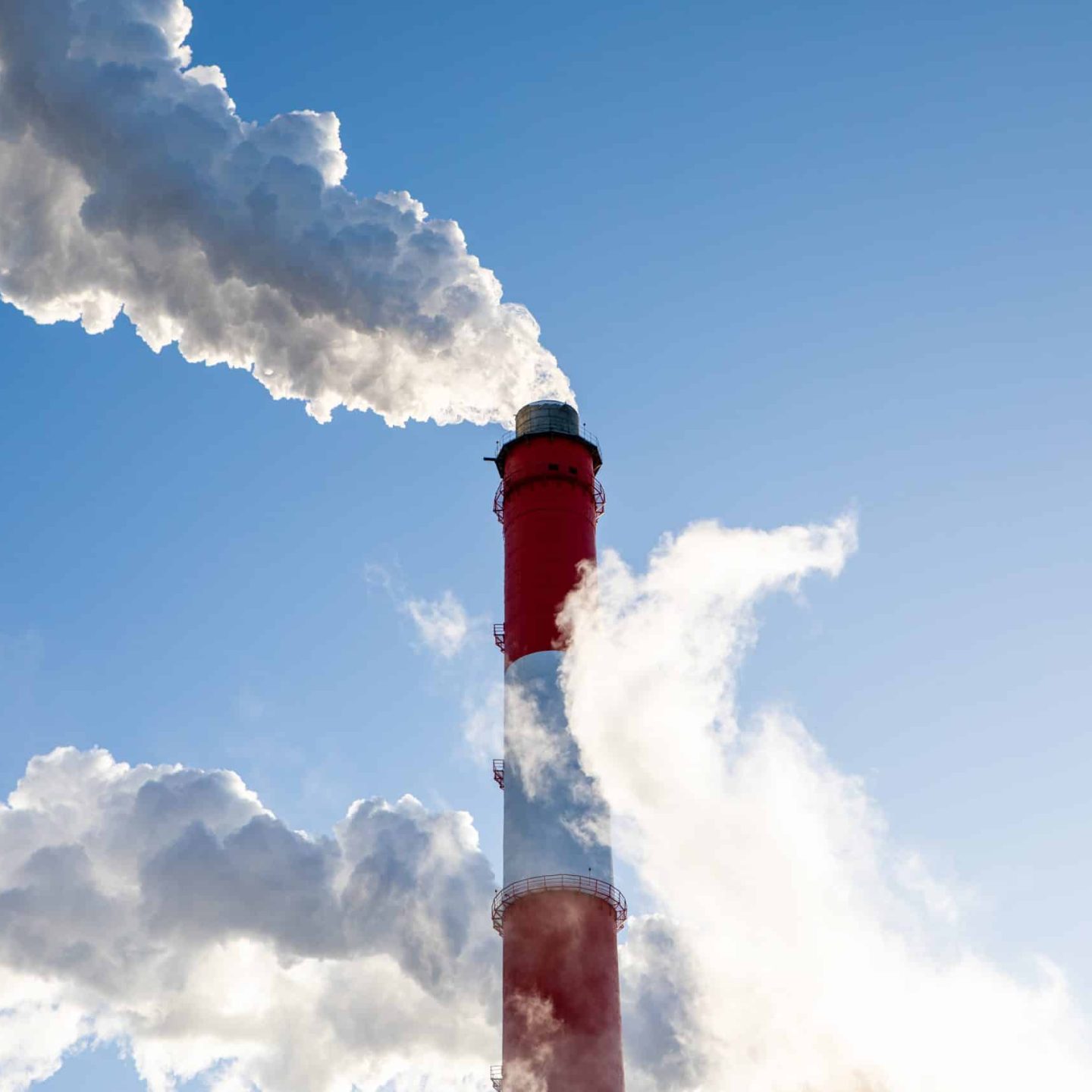 A tall, red-and-white industrial smokestack emits thick white smoke against a clear blue sky, indicating active production or energy generation. Dense clouds of smoke obscure parts of the stack as they disperse into the atmosphere—a typical scene depicted in an Aluminium Sector Factsheet.