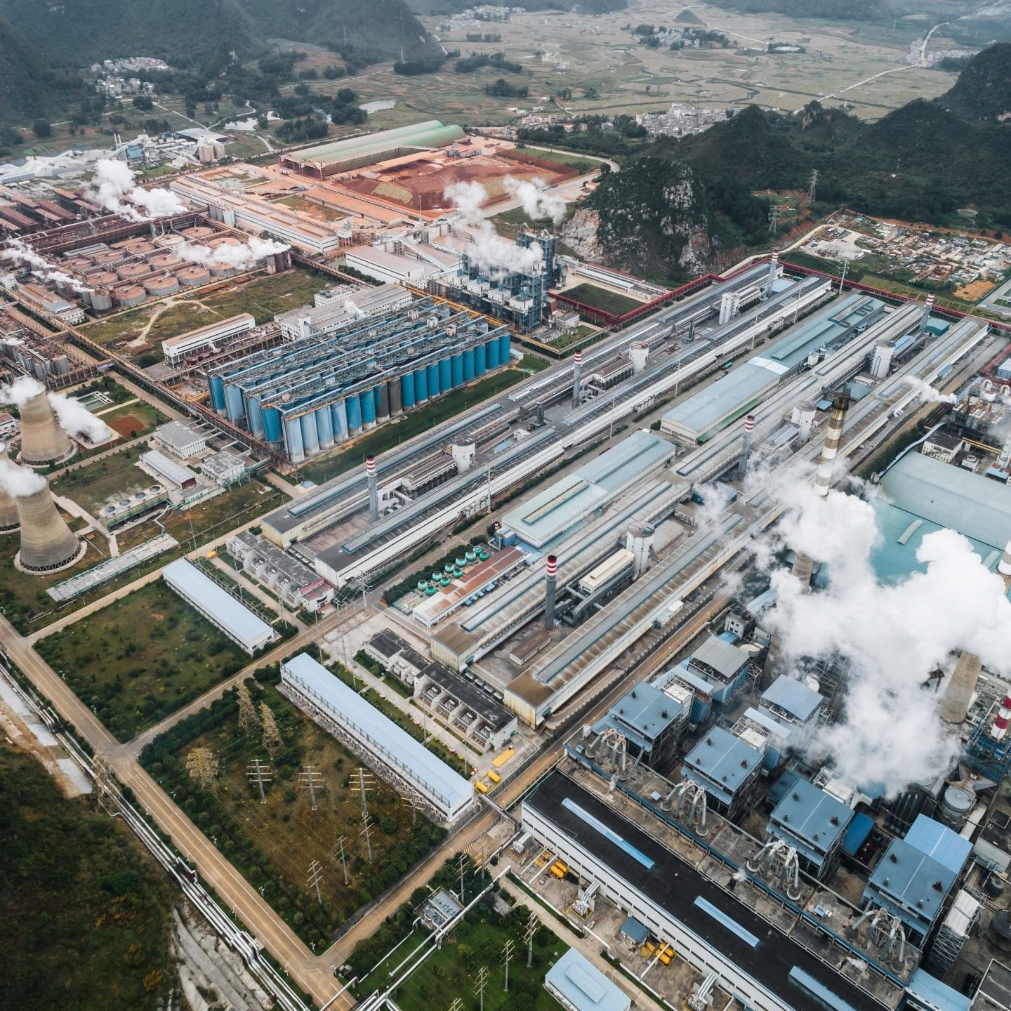 Aerial view of an industrial complex featuring multiple large factories with warehouses, chimneys emitting steam or smoke, and several cooling towers. The surrounding area includes roads, smaller buildings, and hilly terrain in the background—highlighting the challenges and opportunities for sustainable development.