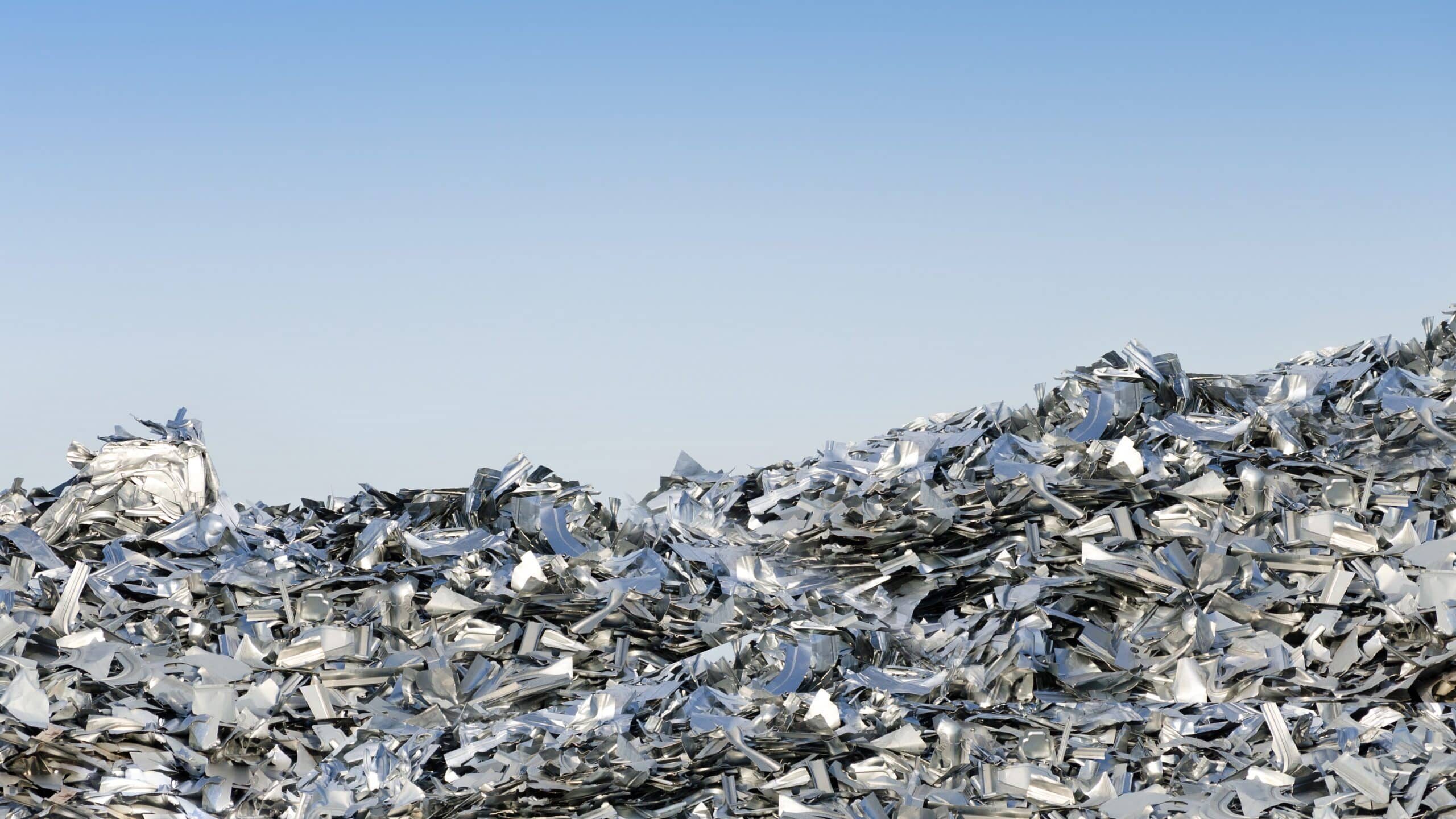 A vast pile of crushed and flattened metallic scrap pieces, primarily for recycling aluminium, stretches into the horizon under a clear blue sky. The metal shards reflect light, creating a shimmering effect across the heap of debris.