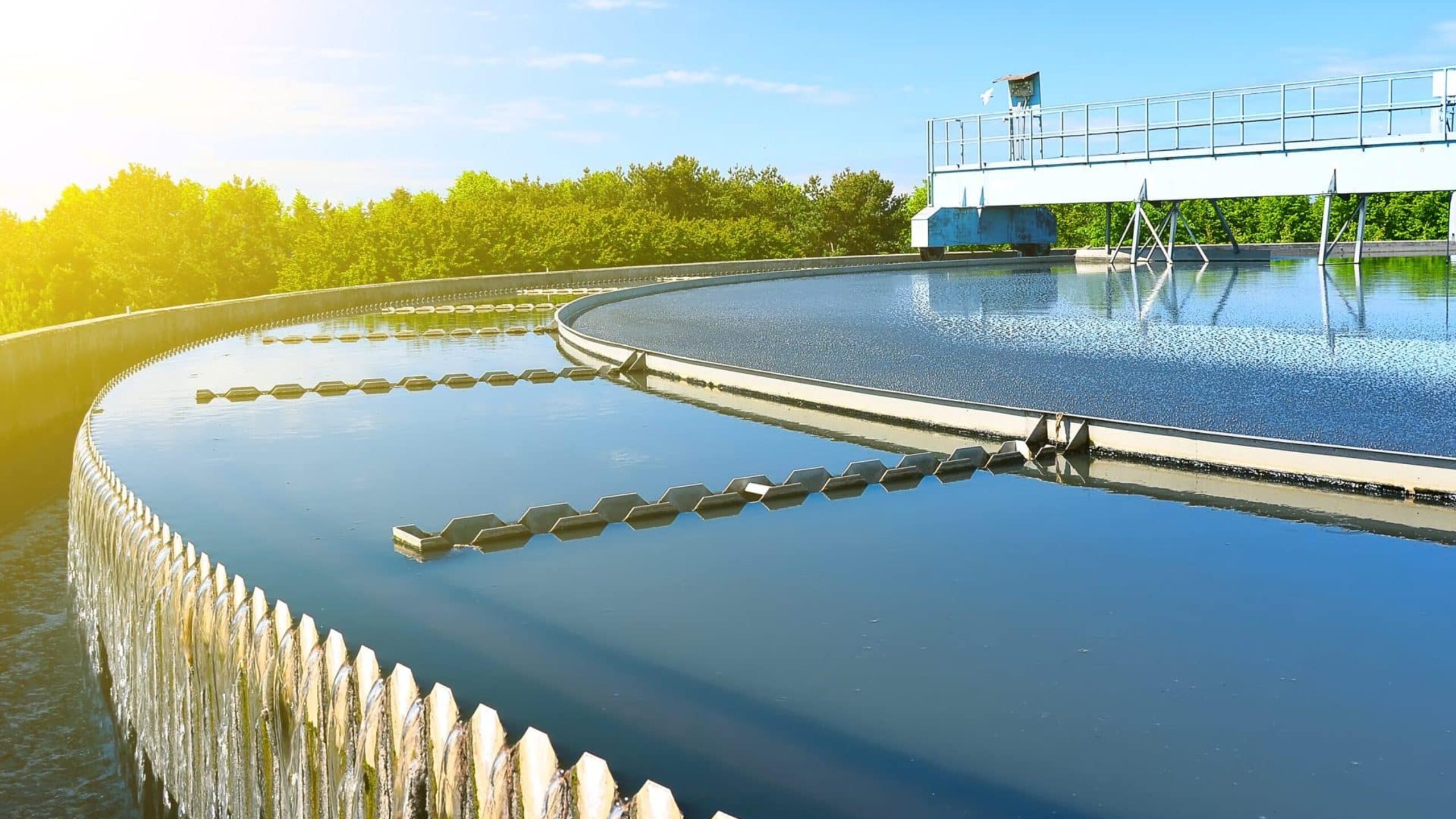 A large circular water treatment facility with a bright blue sky and lush green trees in the background. Clear water flows over the edge of the stepped aluminium structure, reflecting sunlight and creating a serene scene. A walkway with railings spans across the basin.