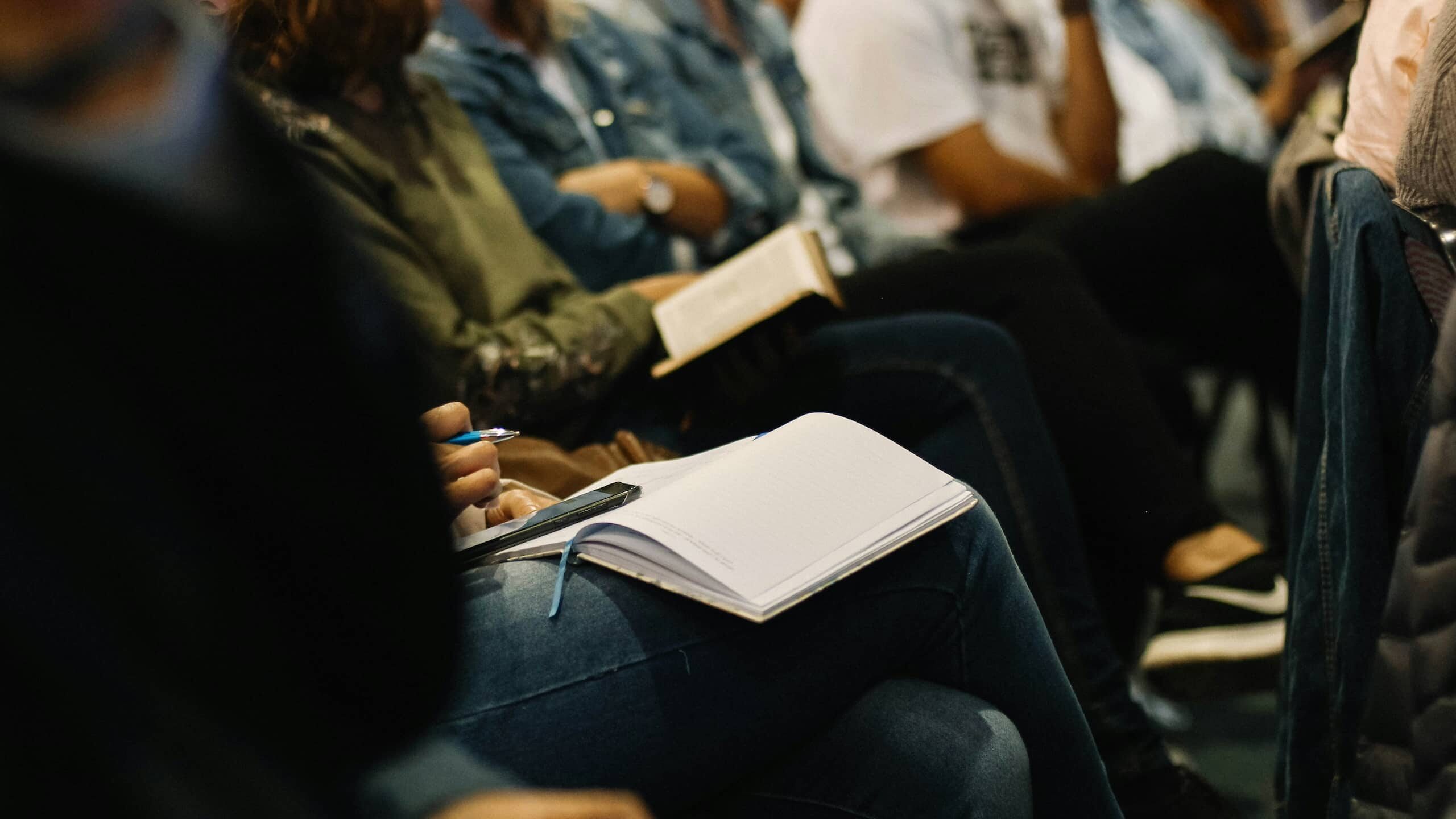 People are seated in a classroom or conference setting, attentively taking notes in notebooks and holding books. The image focuses on their hands and notebooks, emphasizing engagement and learning, symbolizing the foundation of skills needed for the aluminium 2030 industry transformation.