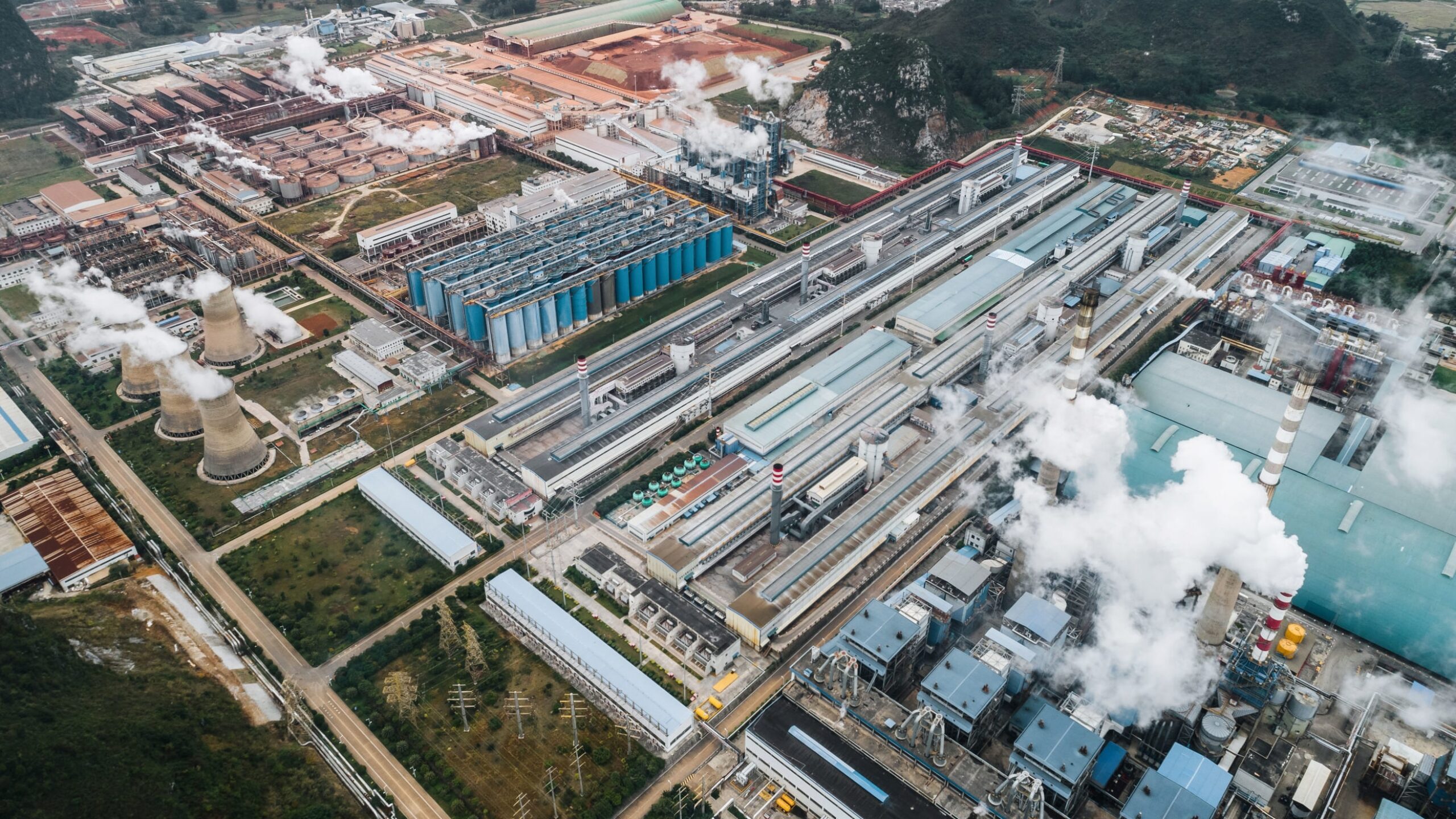 Aerial view of an industrial complex with numerous factories, smokestacks emitting steam or smoke, and multiple long, rectangular buildings. Spent pot lining aluminium waste is managed on-site. The complex is surrounded by greenery and mountainous terrain in the background. Roads and pathways are visible throughout.