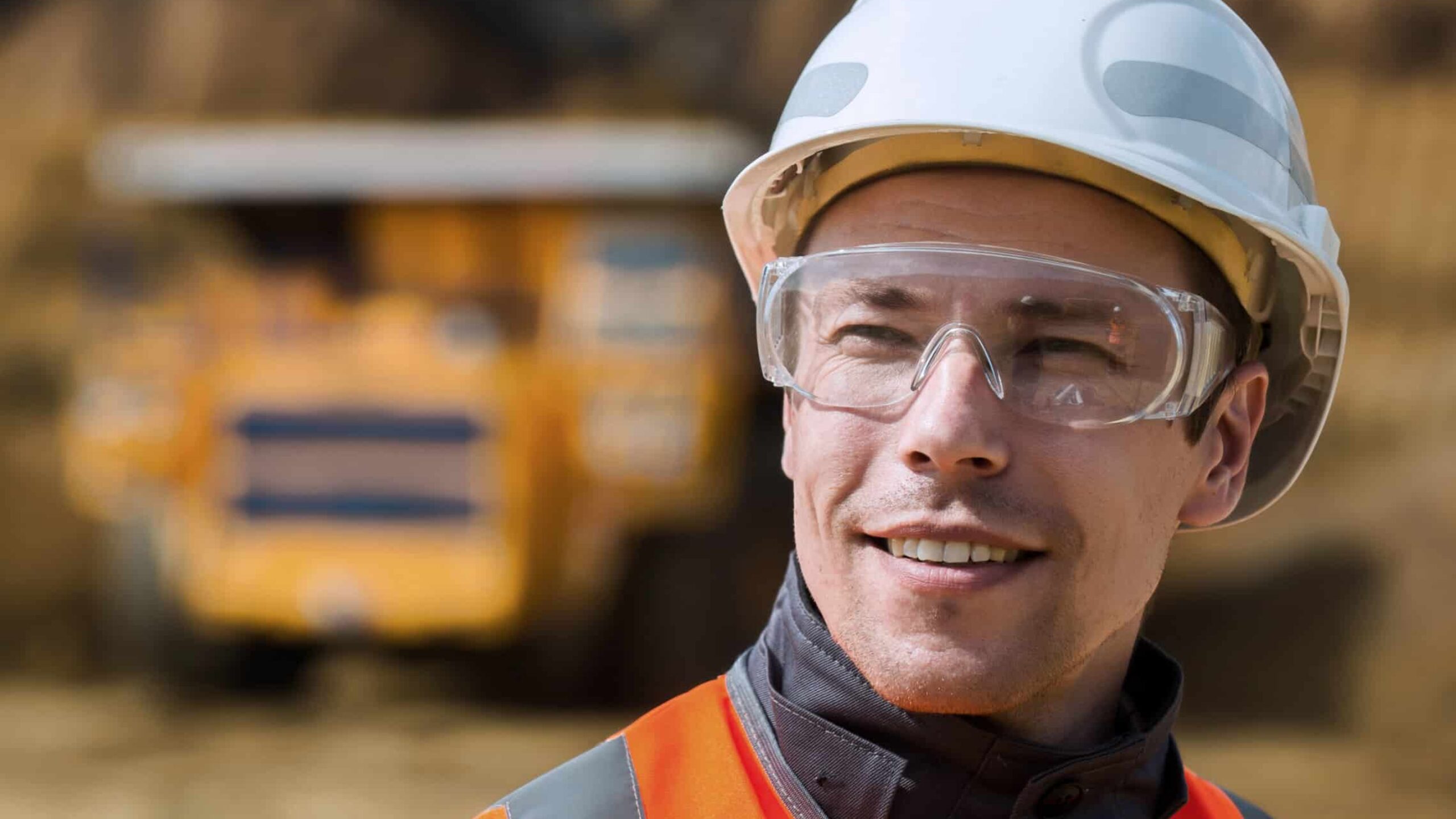 A construction worker wearing a white hard hat, clear safety goggles, and an orange safety vest is smiling. In the blurred background, amid the shimmer of safety aluminium scaffolding, a large yellow construction vehicle stands out. The setting appears to be an active construction site.