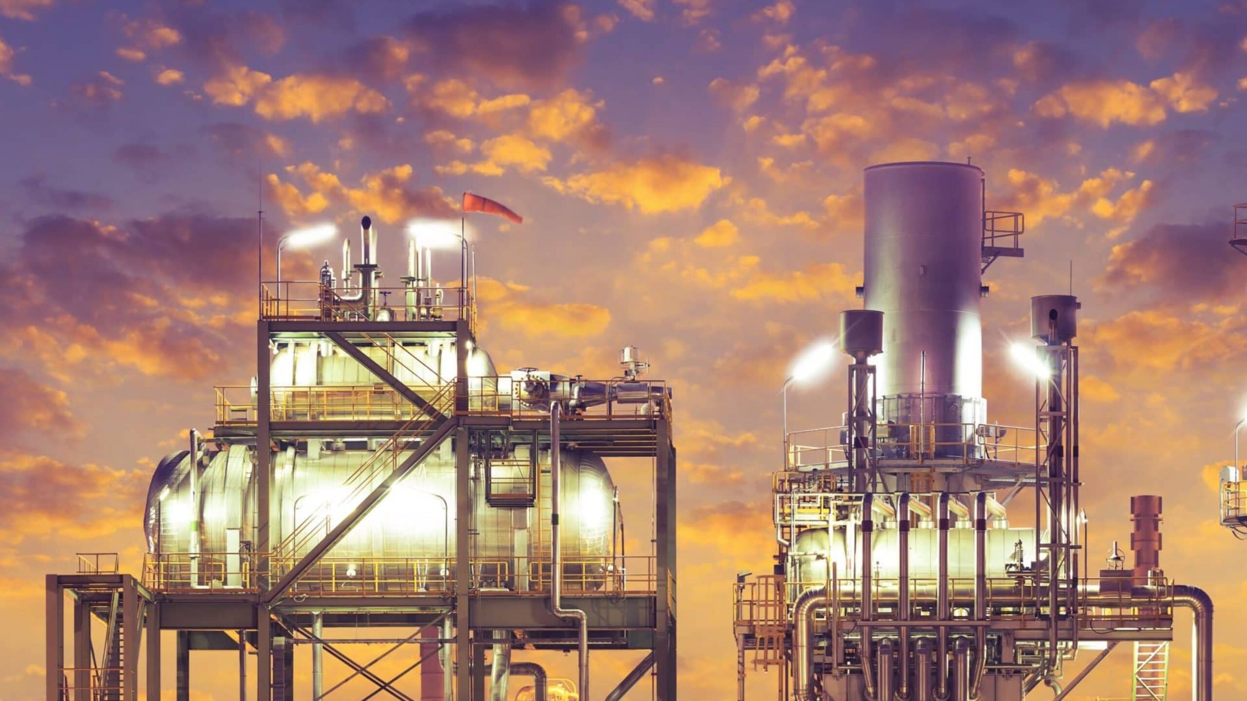 An industrial facility at twilight with illuminated structures and equipment against a backdrop of a colorful, cloud-filled sky. The energy-driven machinery and aluminium pipelines are detailed and prominently lit, contrasting with the warm hues of the sunset.